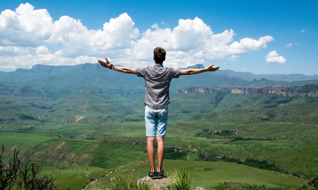 A man stands triumphantly with arms open, overlooking a scenic mountain range.